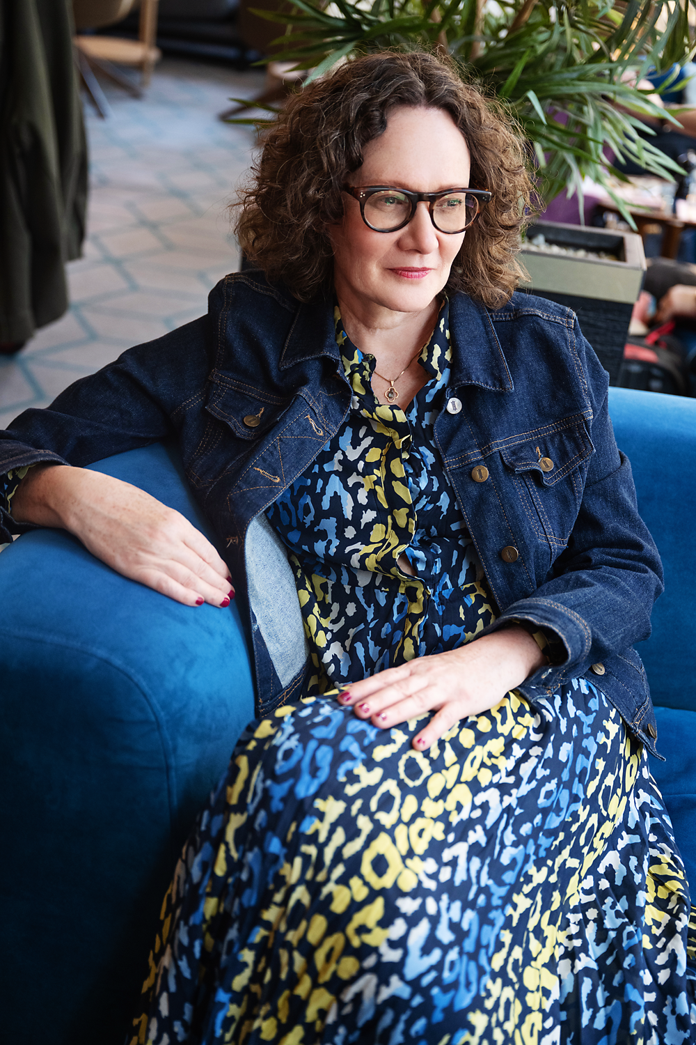 Emma Marinos wearing a denim jacket over a colorful animal print dress, sitting on a blue couch in a public indoor space, with plants and people visible in the background.