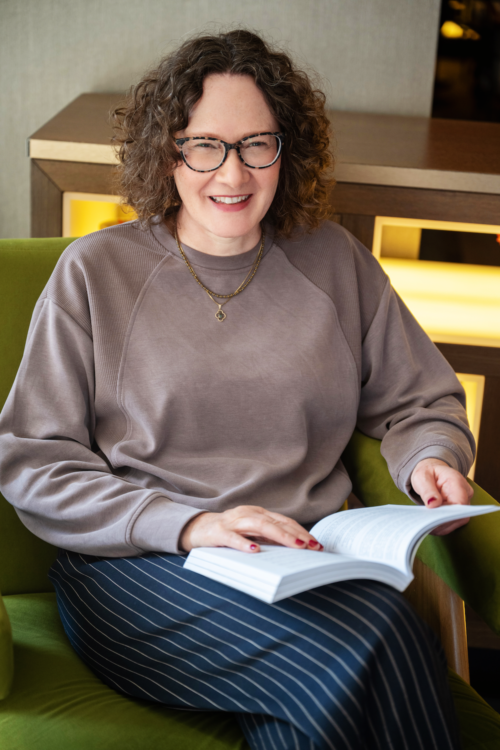 Emma Marinos sitting on a green chair, smiling and reading a book. She is wearing a gray sweatshirt, striped pants, and layered necklaces.