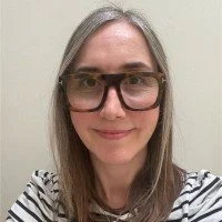 A woman with straight, light brown hair wearing glasses and a black-and-white striped shirt, smiling in front of a plain light-colored wall.