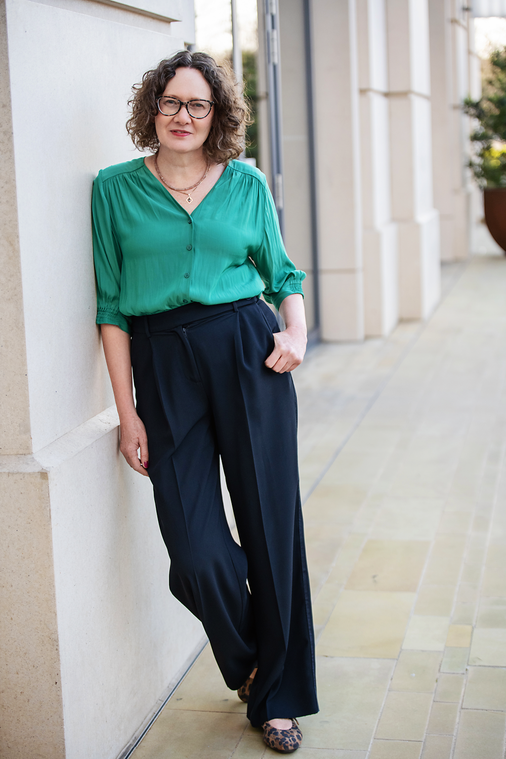 Emma Marinos wearing a green blouse and dark pants, leaning against a wall outdoors.