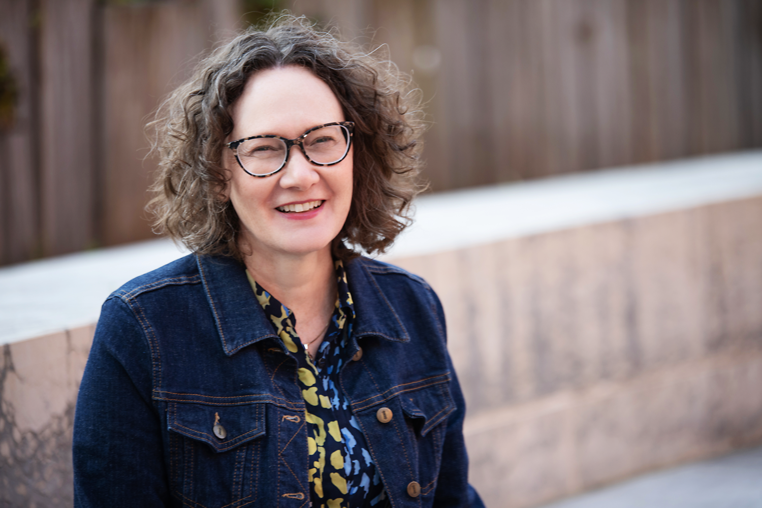 Emma Marinos in a denim jacket smiling outdoors with a wooden fence in the background.