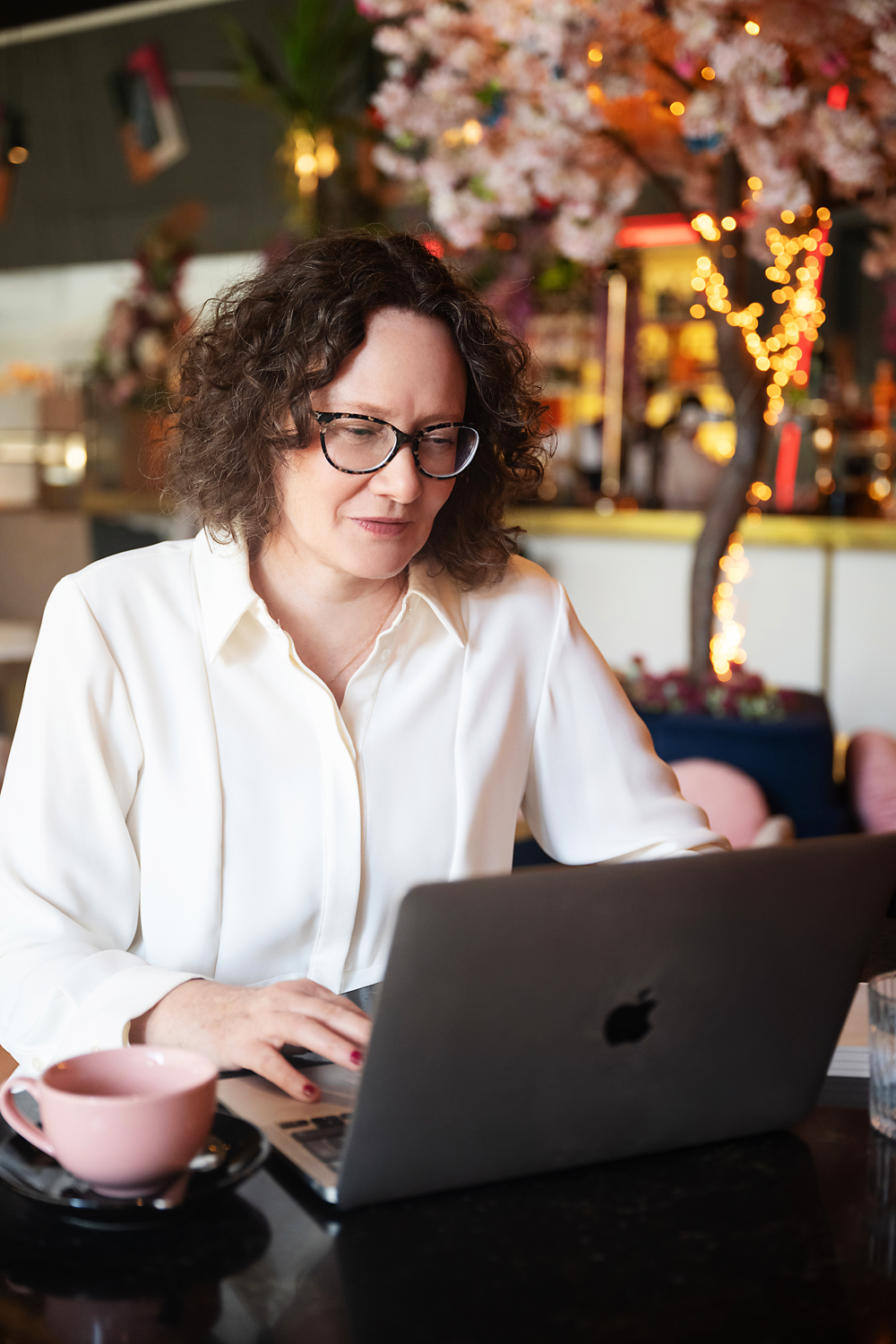 Emma Marinos working on a laptop in a cafe with pink blossoms and string lights in the background.