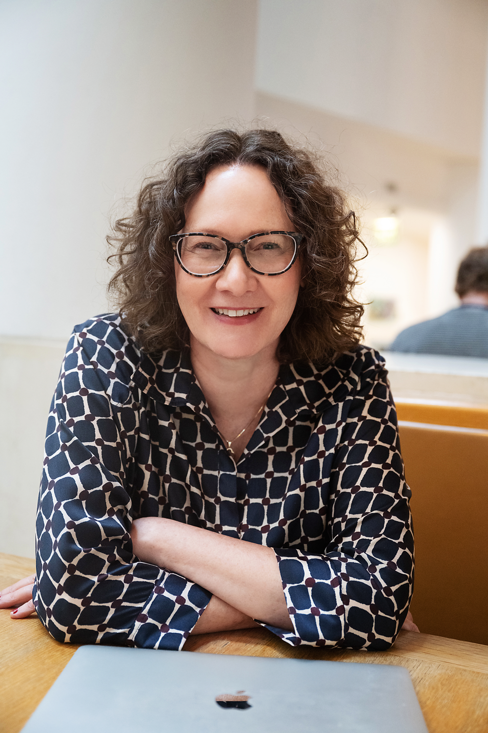 Emma Marinos wearing glasses and a patterned blouse, smiling and sitting at a wooden table with a closed laptop in front of her