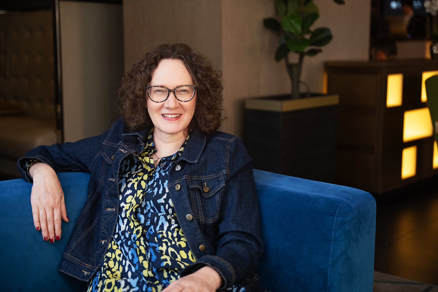 Emma Marinos sitting on a blue couch, smiling at the camera in a modern indoor setting with a decorative lighted cabinet and potted plant in the background.