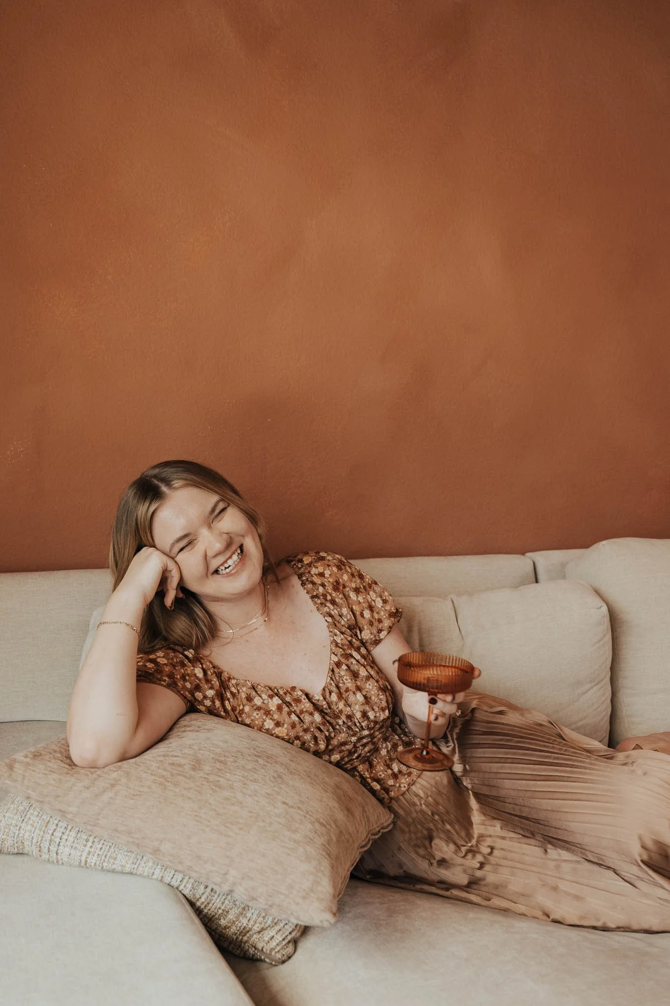 Aliya Mooney, founder and sole creative of Hearthstone Creative, sits on a couch, smiling and holding a fun drinking glass.