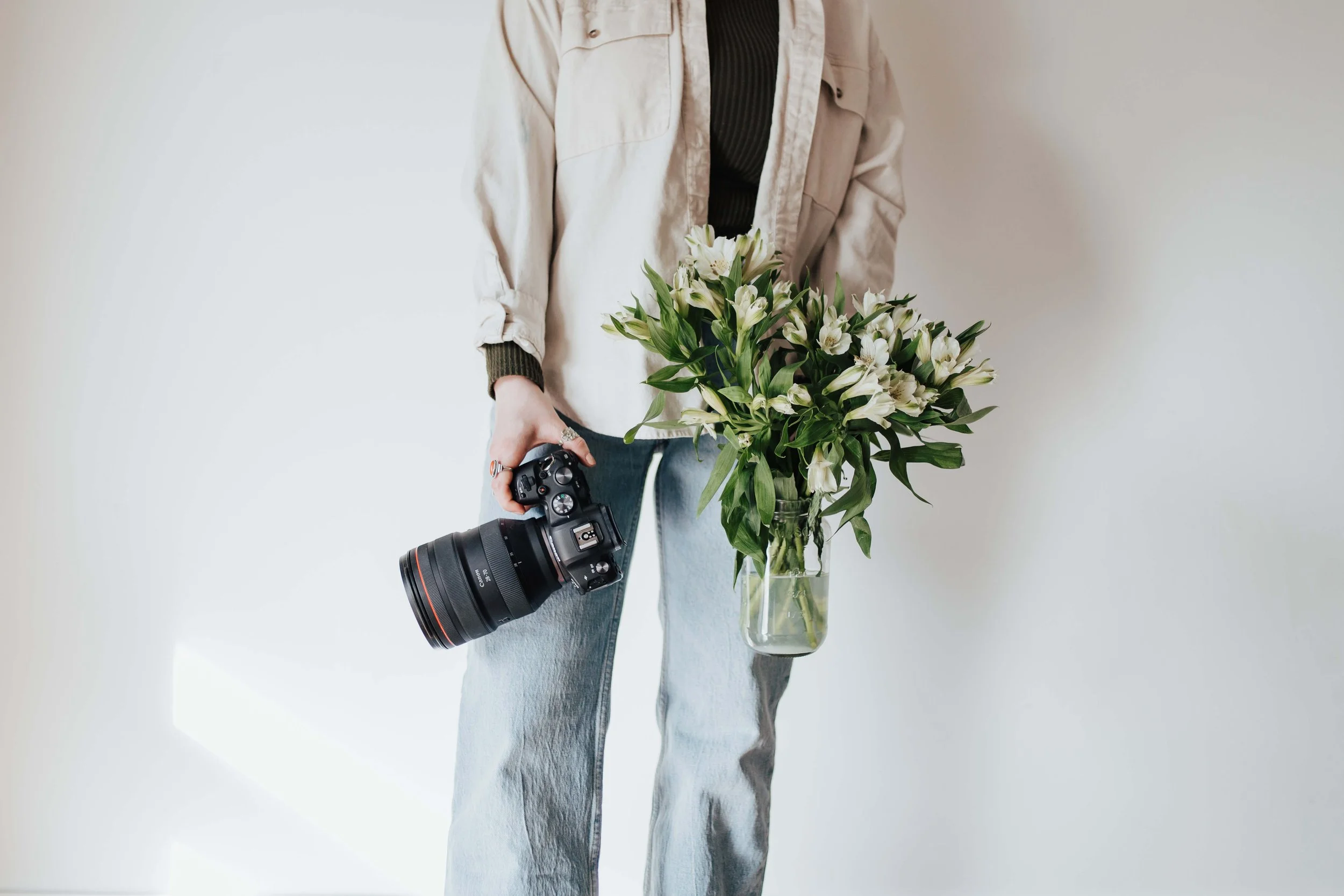 A wedding photographer holds a glass mason jar filled with white flowers in one hand and her camera in the other in front of a white background.