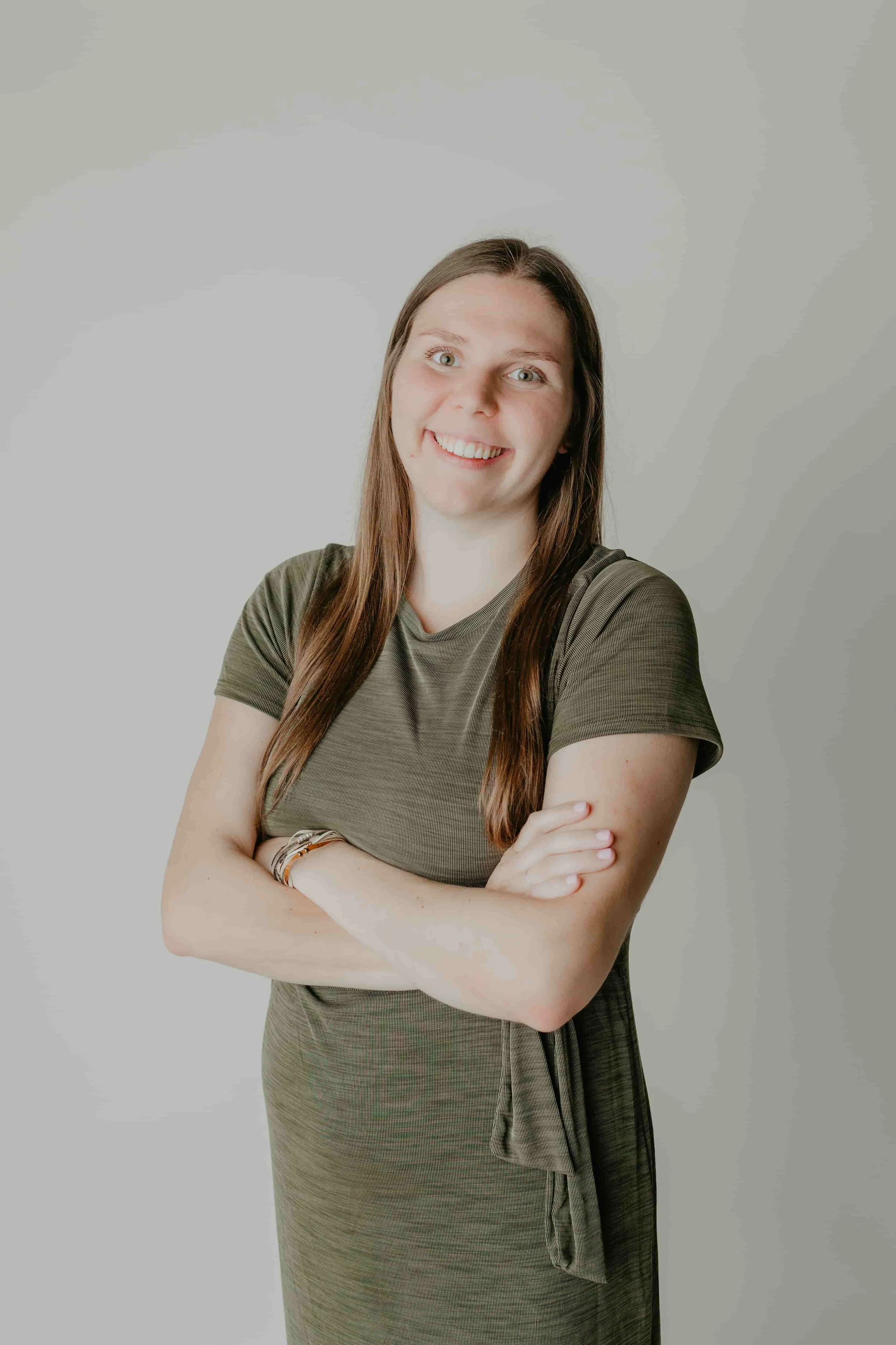 A young woman with long brown hair, wearing a green dress, smiling with arms crossed in front of a plain light-colored background.