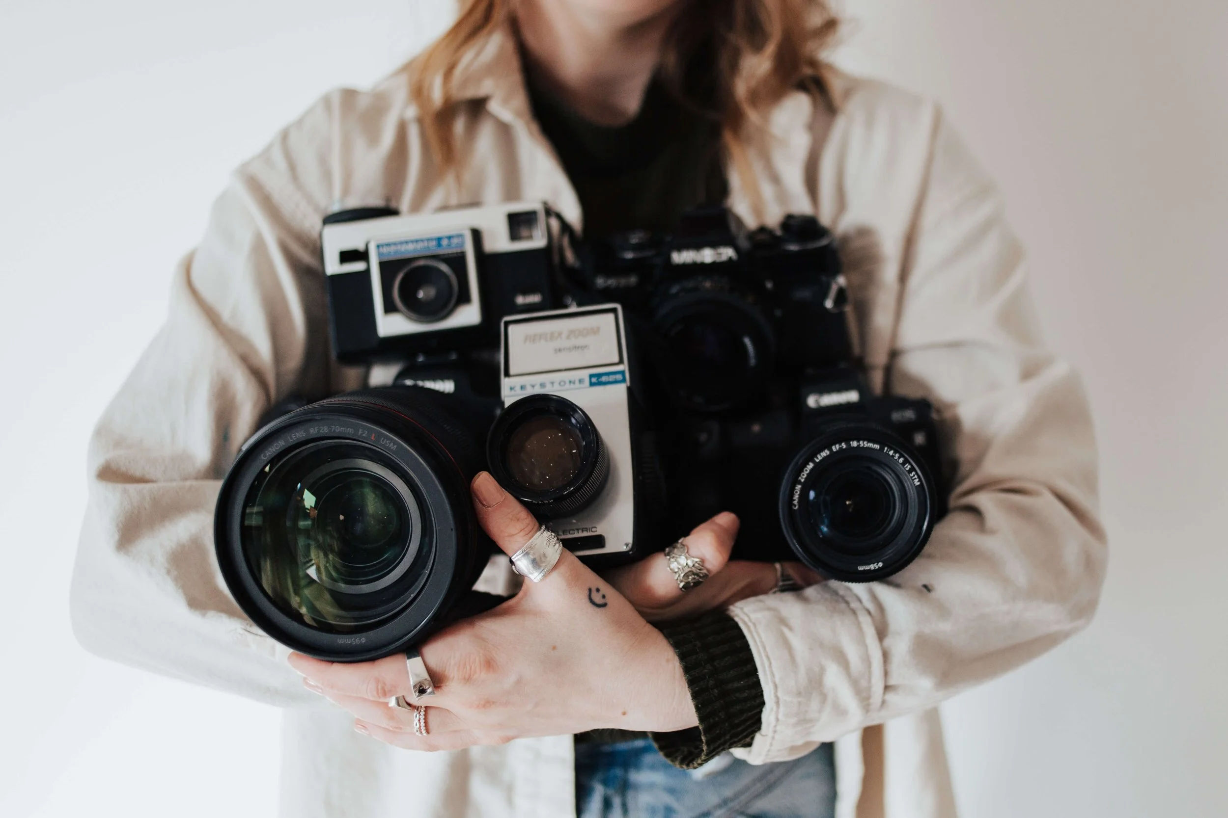 Person holding multiple cameras, including a large camera lens, an instant camera, and a digital camera, wearing rings and a beige jacket.