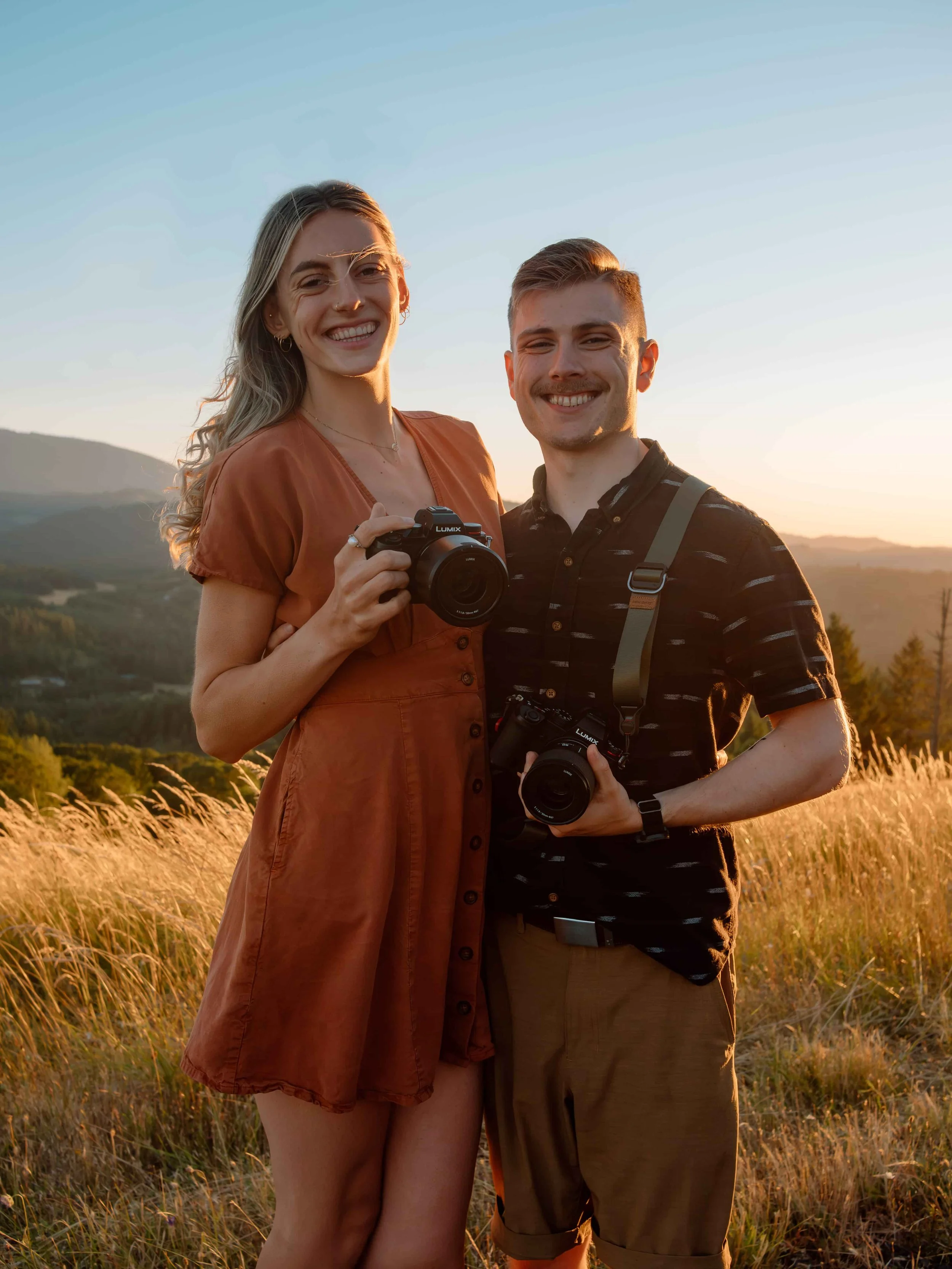 A photographer and videographer stand in a golden field holding their cameras.