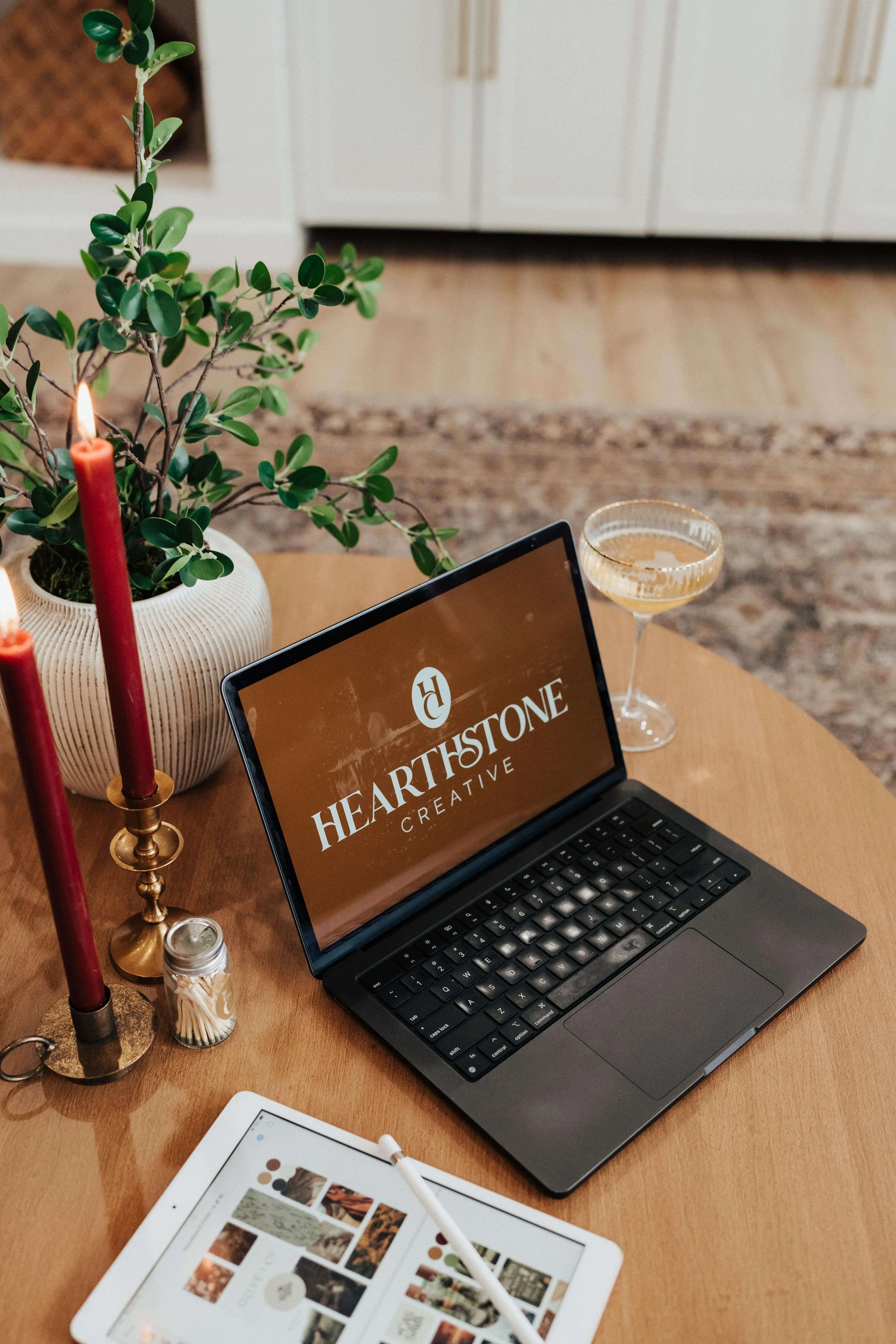 A laptop displaying the Hearthstone Creative logo sits on a table next to a tablet with a mood board open, two lit candles, and a glass of sparkling cider.