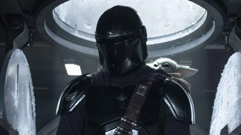 Upper body shot of Din Djarin with Grogu peeking over his shoulder. Djarin is standing with the interior of a generic-looking ship in the background with some frosty detailing.