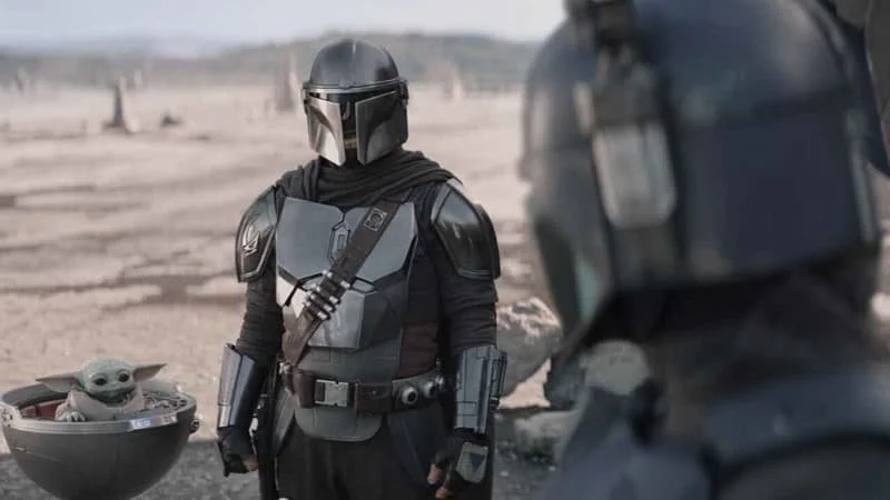 Season three promo image for The Mandalorian, Din Djarin and Grogu in his pram facing Paz Vizla, who is slightly out of focus with his back facing the camera.  Image Source: IMDb
