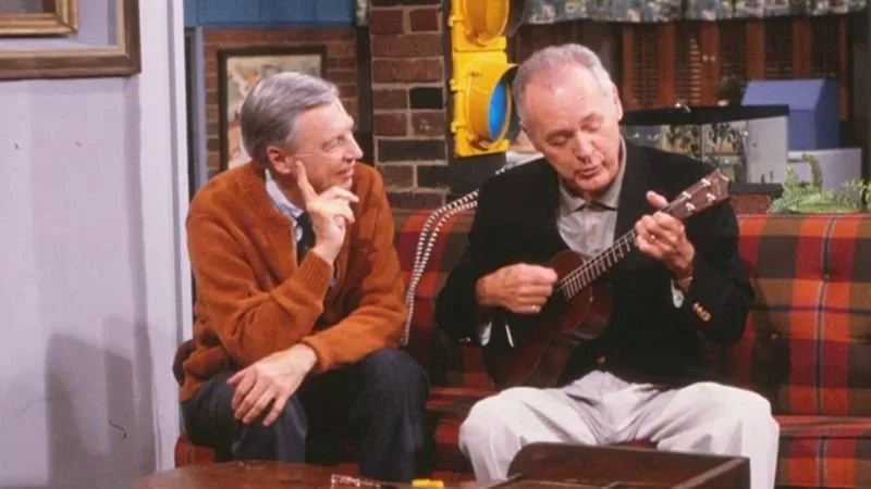 This image features Fred Rogers wearing an orange cardigan sweater alongside Joe Negri holding a ukulele.
