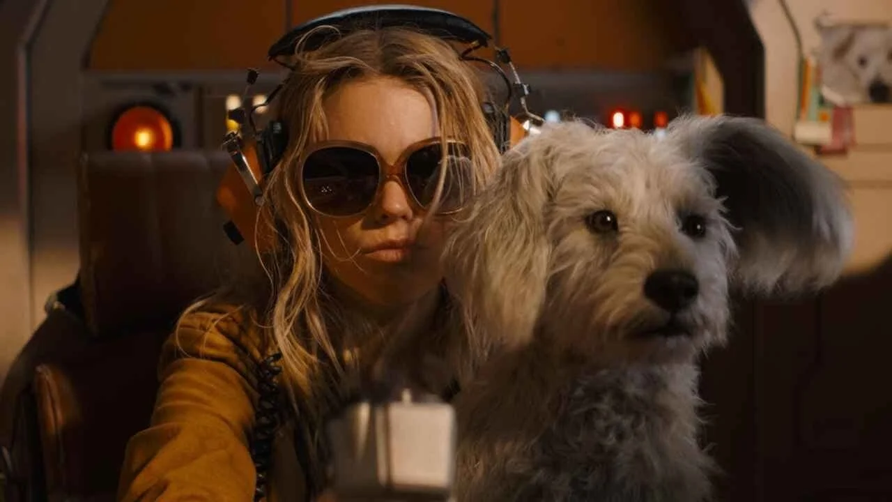Kara and Krypto in a spaceship cockpit facing a light source like a window, Krypto is sitting on Kara's lap. Kara is wearing large orange sunglasses and orange headphones. Image Source: IMDb