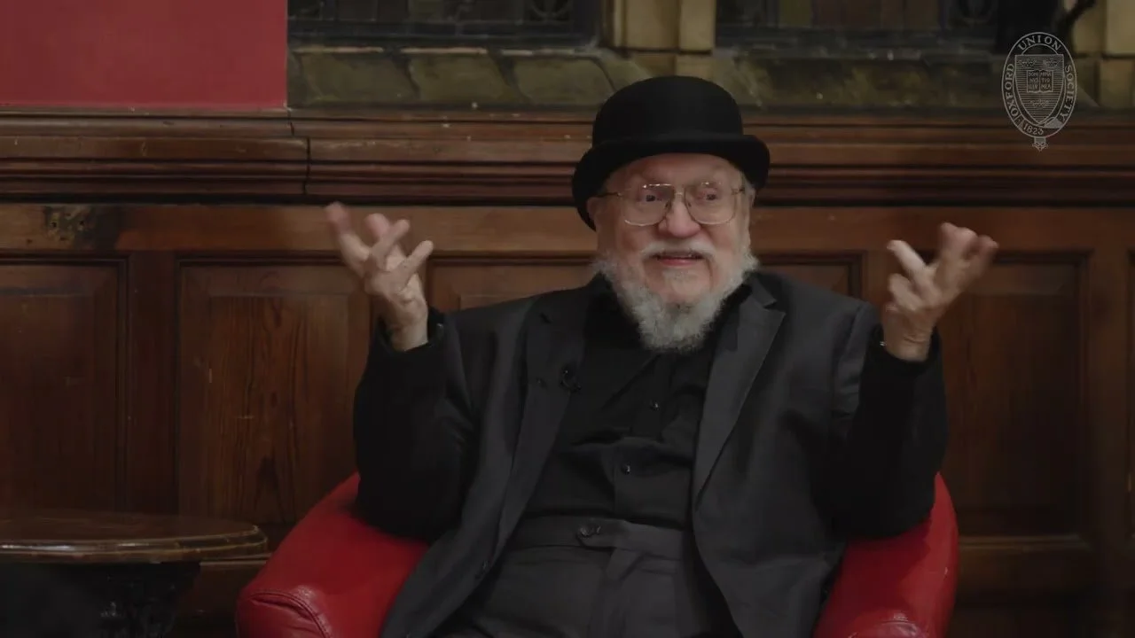 Martin in a red chair raising his hands  Image Source: Not A Blog - George R.R. Martin