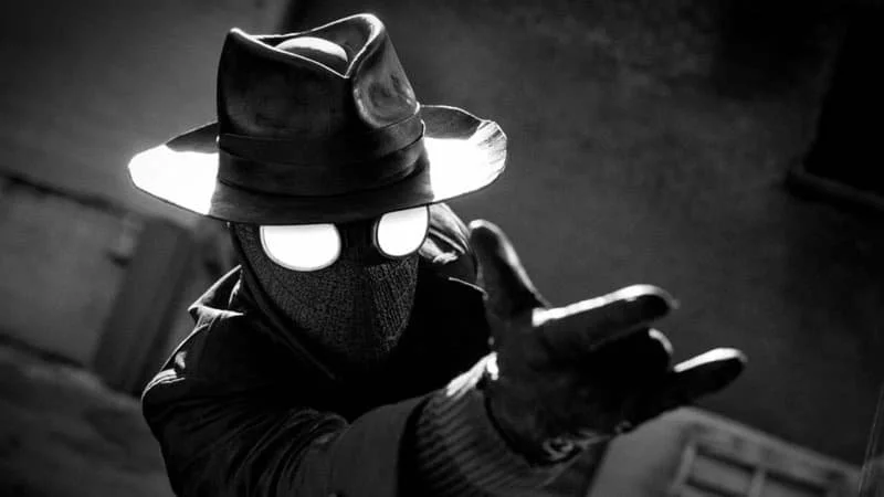 Nic Cage’s Spider-Noir; image is black and white, actor is in a Spider-Man suit with a fedora and a rain jacket-esque coat, holding his hand in the classic web-slinging way
