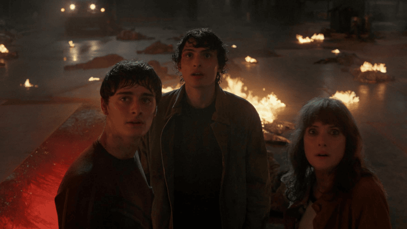 Will, Mike, and Joyce look at something happening in front of them as flaming bodies burn behind them.