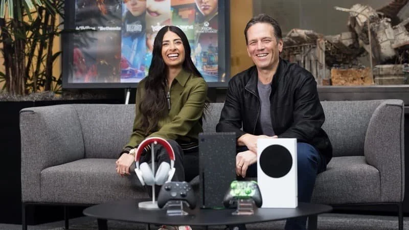 Image depicts former Xbox head Phil Spencer and new XBOX head Asha Sharma on a couch behind Xbox Series models and peripherals  Image Source: IGN