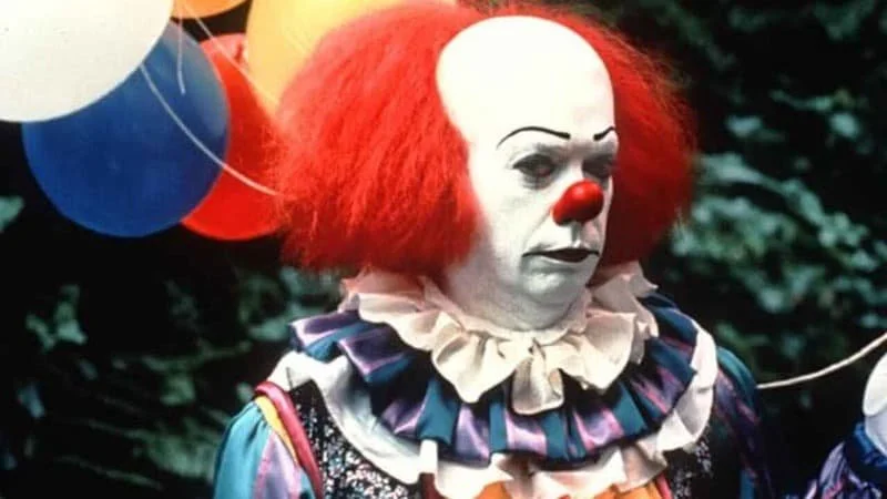 Tim Curry as Pennywise in the 1990 IT miniseries. He is frowning in clown makeup, side-eyeing the camera, and holding a bundle of balloons.  Image Source: IMDb