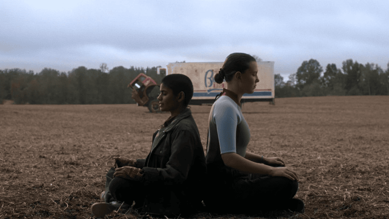 Eleven and Kali sit cross-legged on a field back to back.
