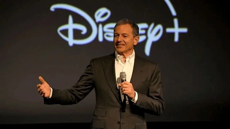 Bob Iger giving a talk, wearing a dark suit and holding a microphone. the background is off-black with the Disney+ logo