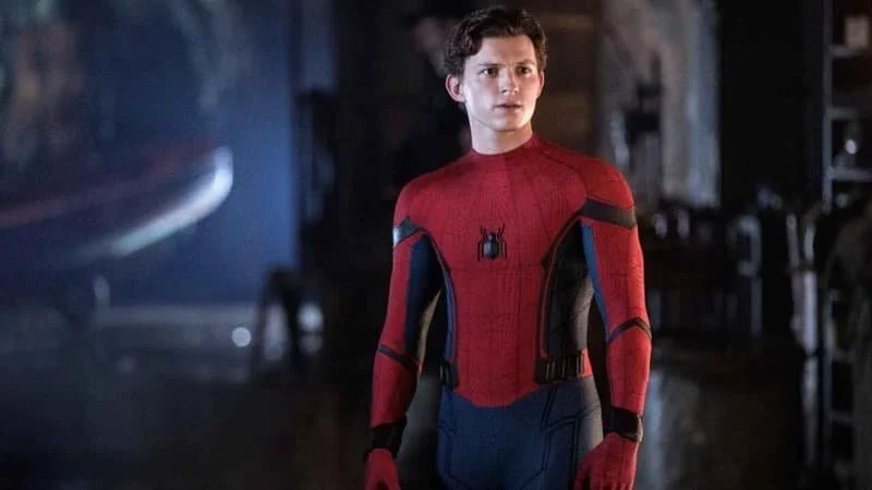 Tom Holland as Peter Parker in Spider-Man: Far From Home (2019). Holland is standing against a dark background in the Spider-Man suit, mask off and in hand, staring to the side of the camera with a lightly furrowed brow.  Image Source: IMDb
