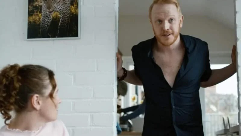 Owain Rhys Davies in A Serial Killer's Guide to Life (2019), standing in a doorway looking down at a woman with the top of his navy blue shirt undone. Image Source: IMDb