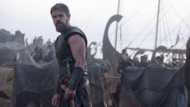 Matt Damon as Odysseus in The Odyssey (2026), character is looking past the camera with a serious expression on his face, the background full of ships, tents, and other people.  Image Source: IMDb