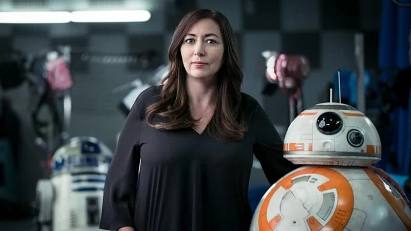 Lynwen Brennan stands in a studio with BB-8 in front and R2-D2 behind.