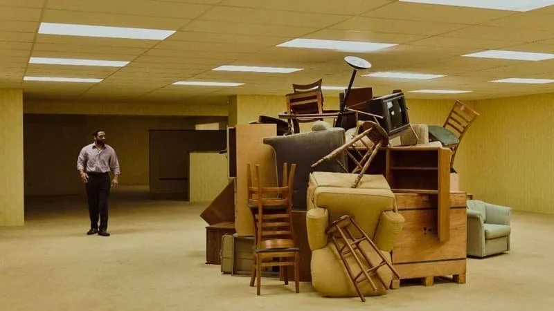 Clark is walking through an area in the Backrooms with a pile of furniture in the center. Clark is looking at the pile while walking.  Image Source: IMDb
