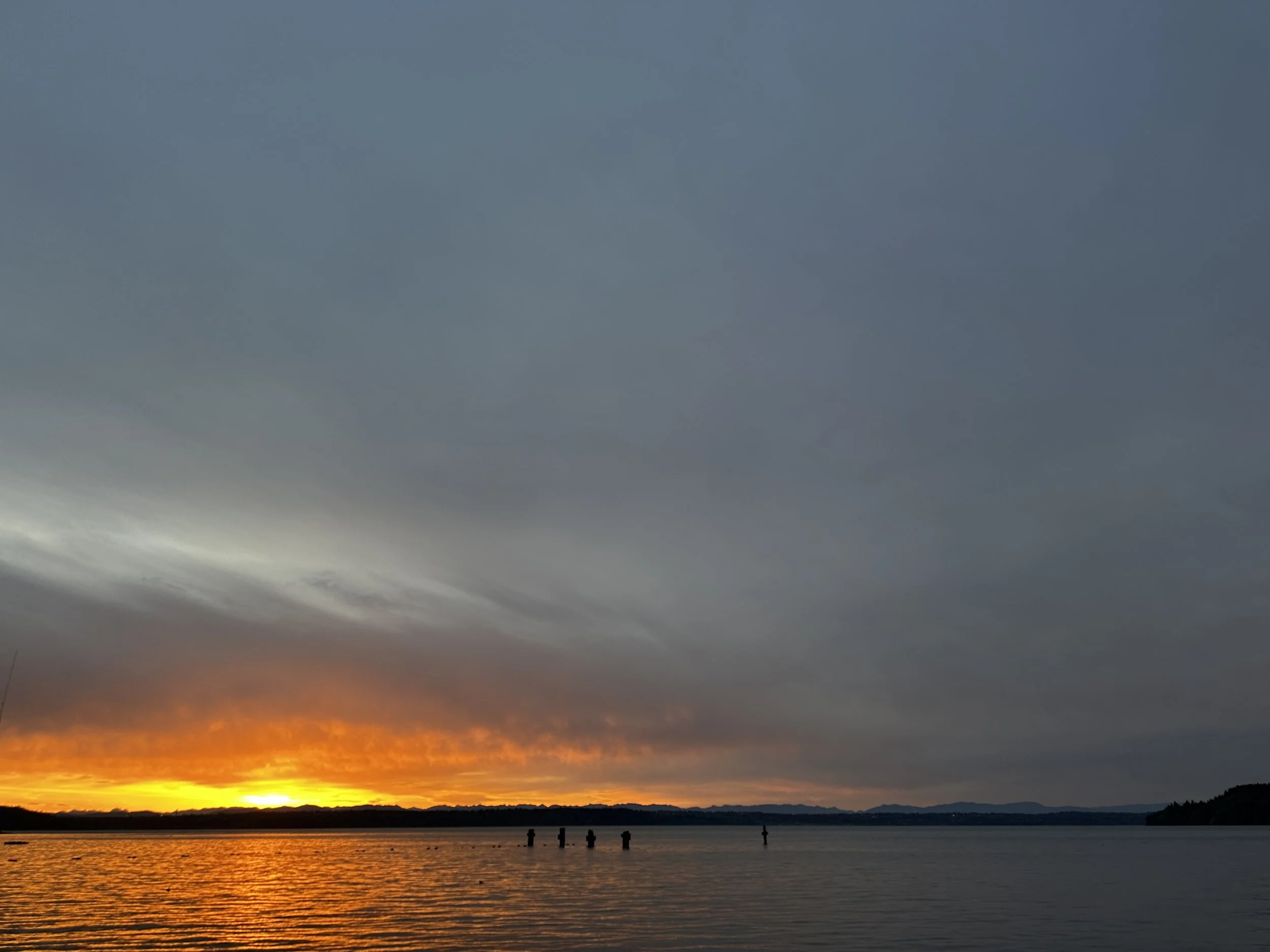 A vibrant yellow sunrise breaks through the gray morning clouds over the water of puget sound, silhoutteing the land.