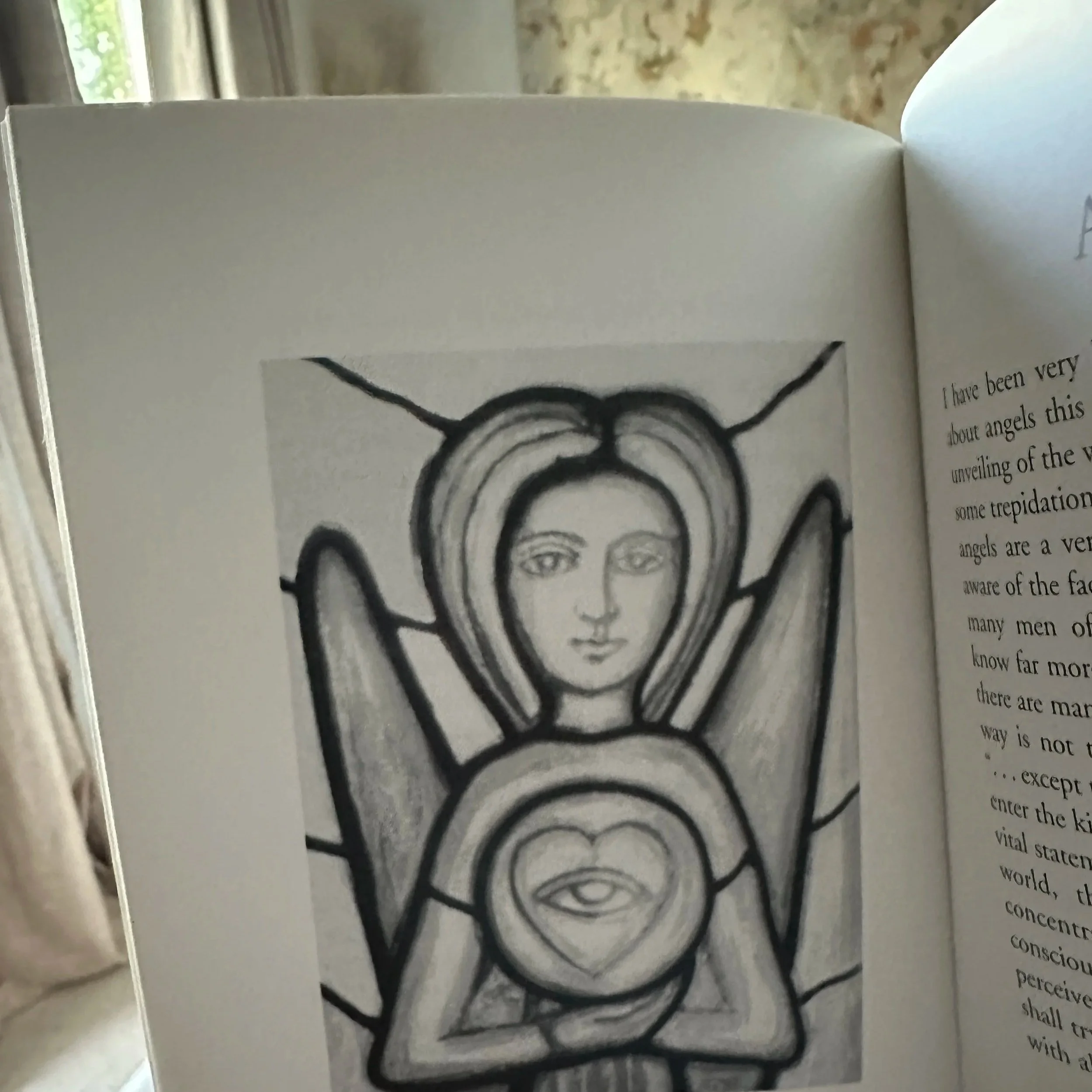 Close-up of an open copy of Angels held in one hand. The left page features a black-and-white illustration of an angel with raised wings and a single eye inside a heart-shaped form at its chest; part of the facing text page is visible.
