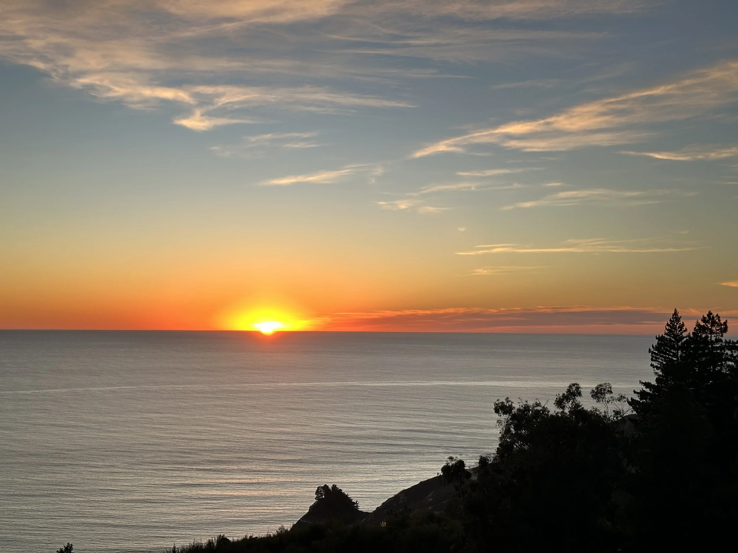 A bright yellow spherical sun rests on the crisp blue oceanic horizon. A diffuse yellow atmosphere extends from the sun. Above the ocean, the sunset gradient starts orange and vertically shifts to light blow. There are gauzy white stripes of clouds.