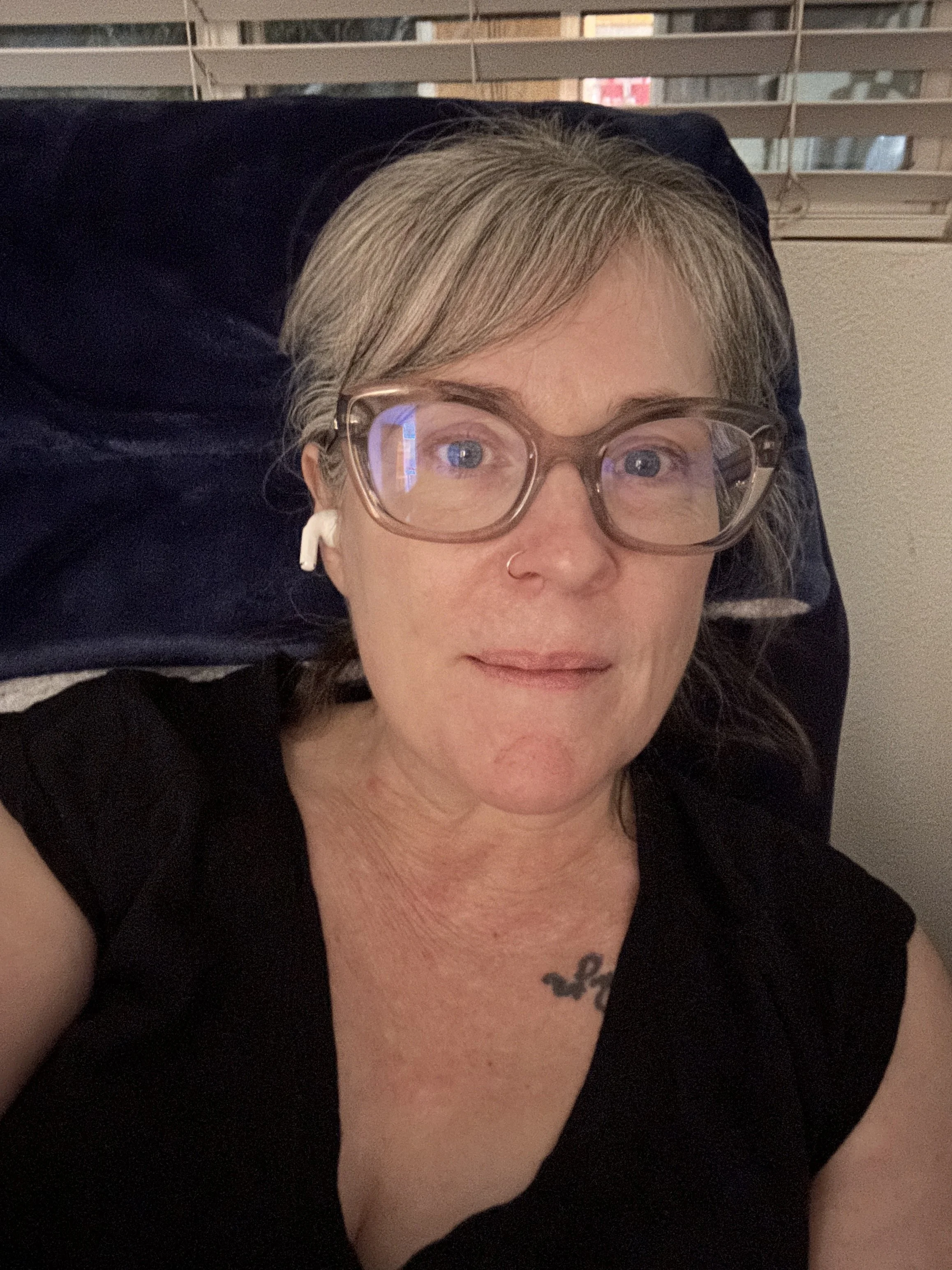 A close-up selfie of a woman with gray-blonde hair and side-swept bangs, translucent champage-colored glasses, a black V-neck shirt with a script tattoo visible on her collar bone sitting in a navy blue recliner in front of a window with blinds.