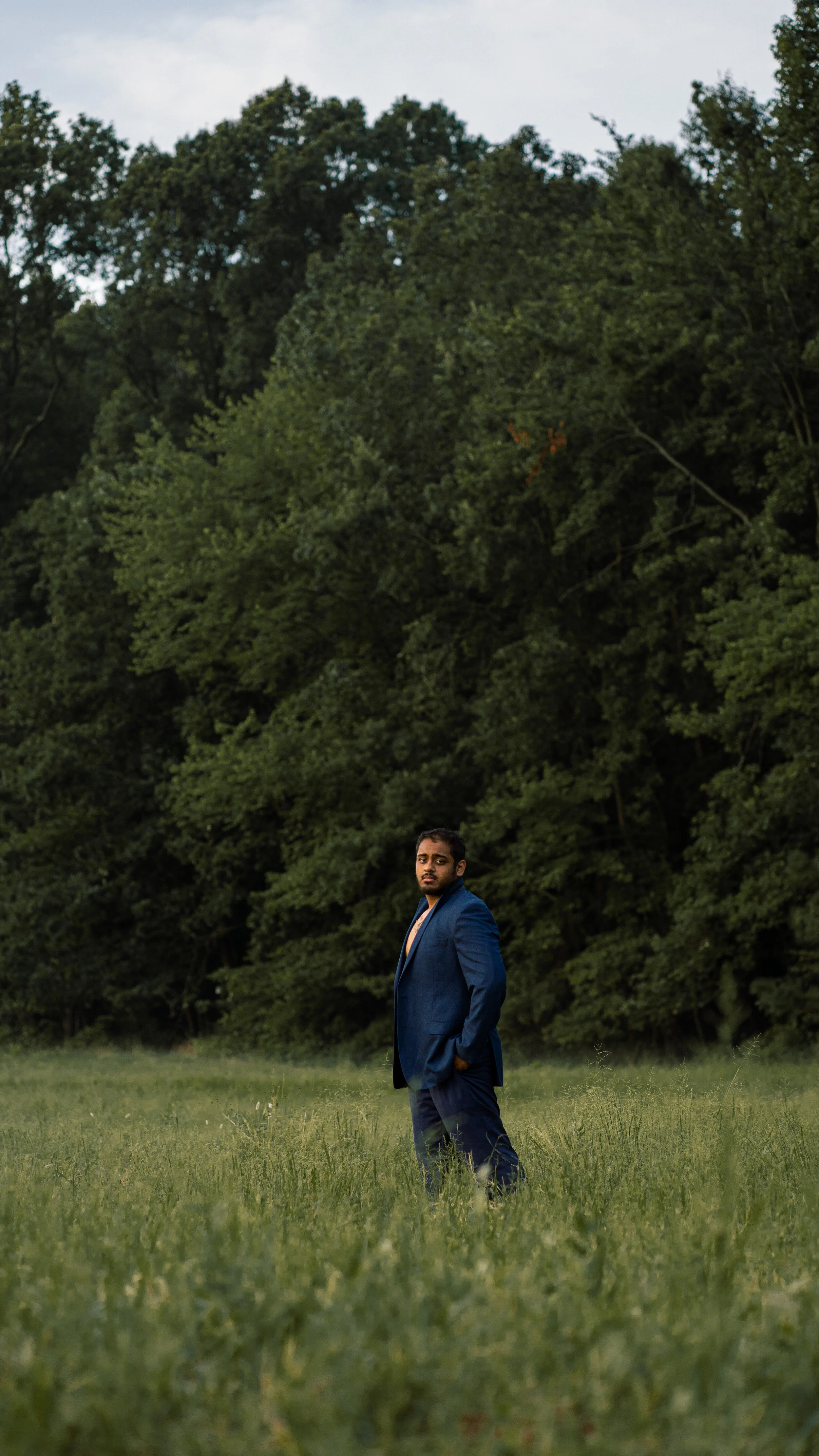Man in a blue suit standing in a grassy field with trees in the background.