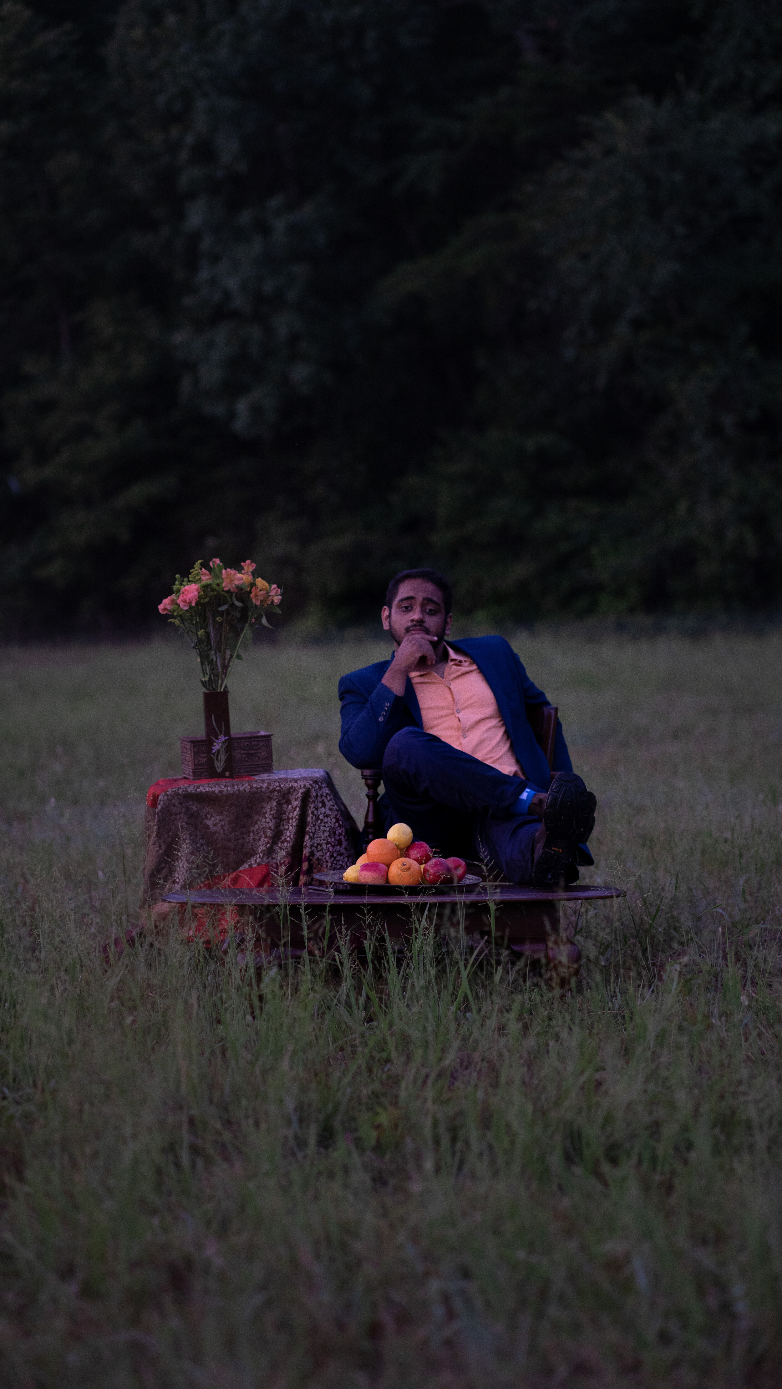 A man dressed in a blue suit and beige shirt sitting on a low platform outdoors during dusk, with a table beside him decorated with flowers and a variety of fruits, in a grassy area with trees in the background.