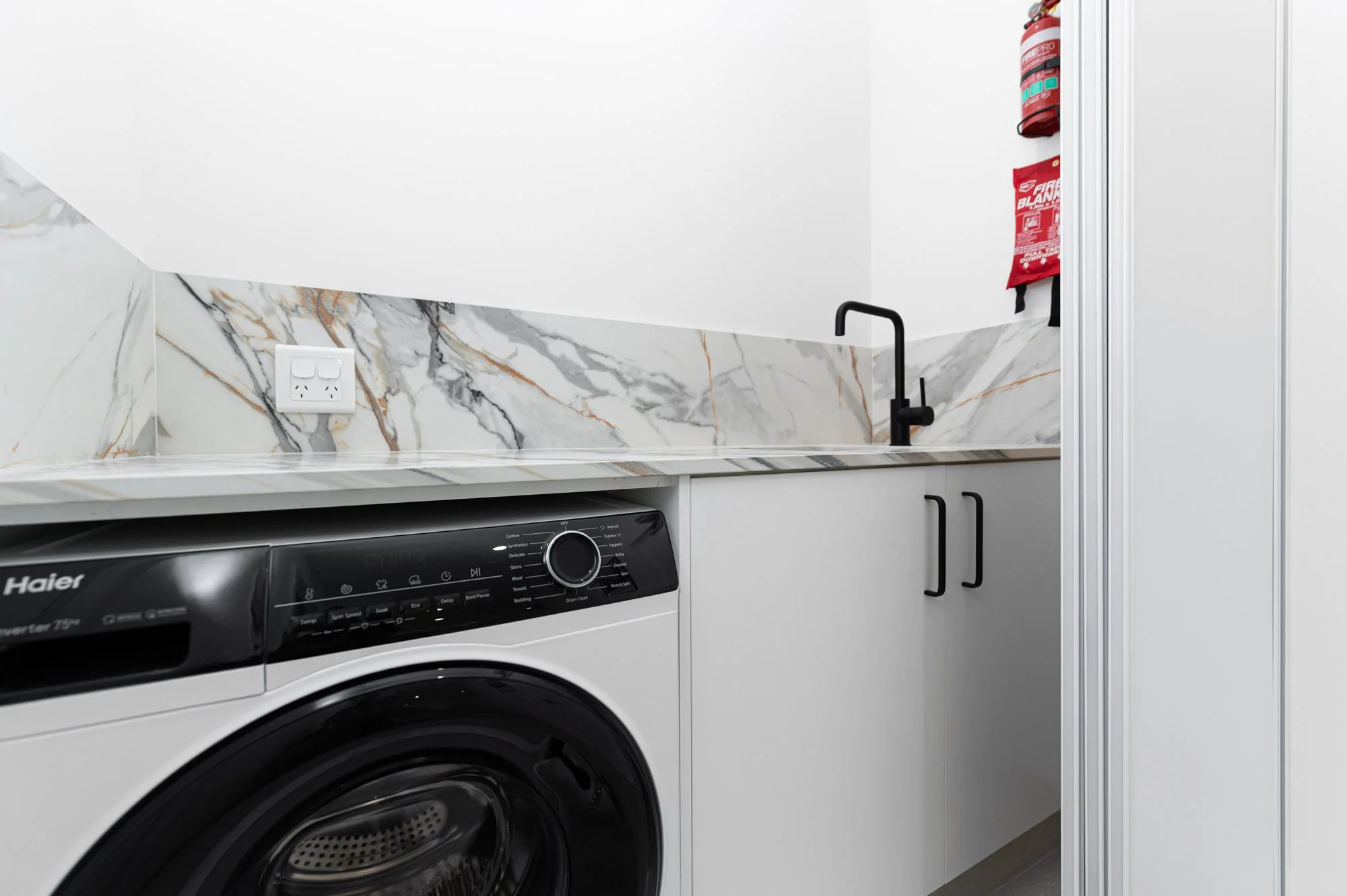 Modern laundry room with black washing machine, marble countertops and backsplash, white cabinetry, and a black faucet.