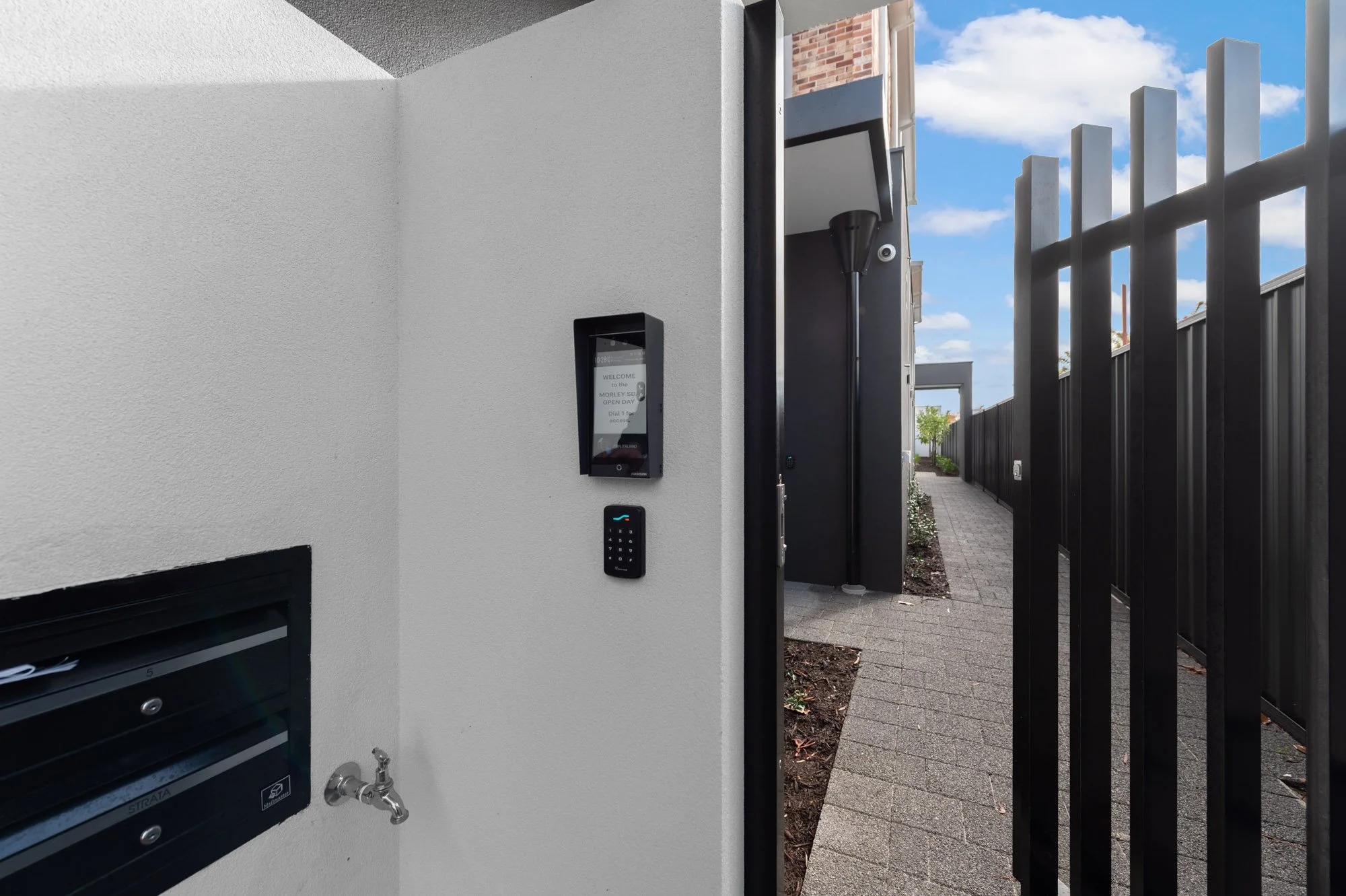 Close-up of a modern house entryway with an intercom system, keypad lock, mailbox, and a black fence along a paved walkway outside.