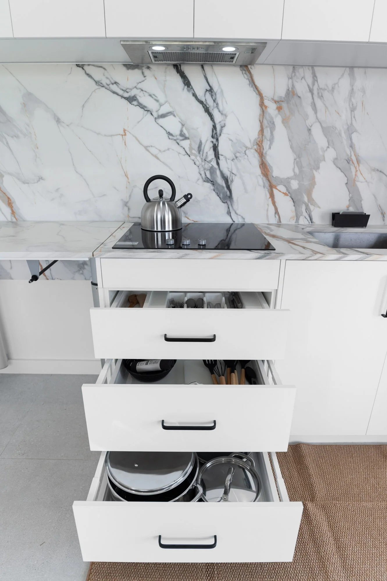 Open kitchen drawers filled with pots, pans, utensils, and kitchen tools, with a marble backsplash and countertop, and a stainless steel kettle on the stovetop.
