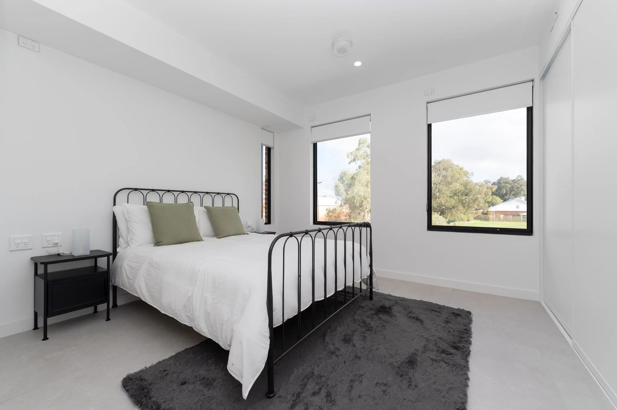 Modern bedroom with white walls, black metal bed frame, white bedding, two green pillows, black side table with lamps, and three large windows showing trees outside.