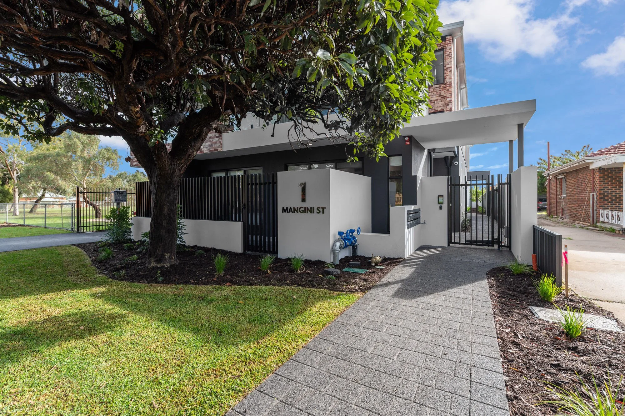 Modern residential building with a white and black exterior, featuring a tree in the front yard, a sidewalk, and a gated entrance with the address 'MANGINI ST' displayed on the wall.