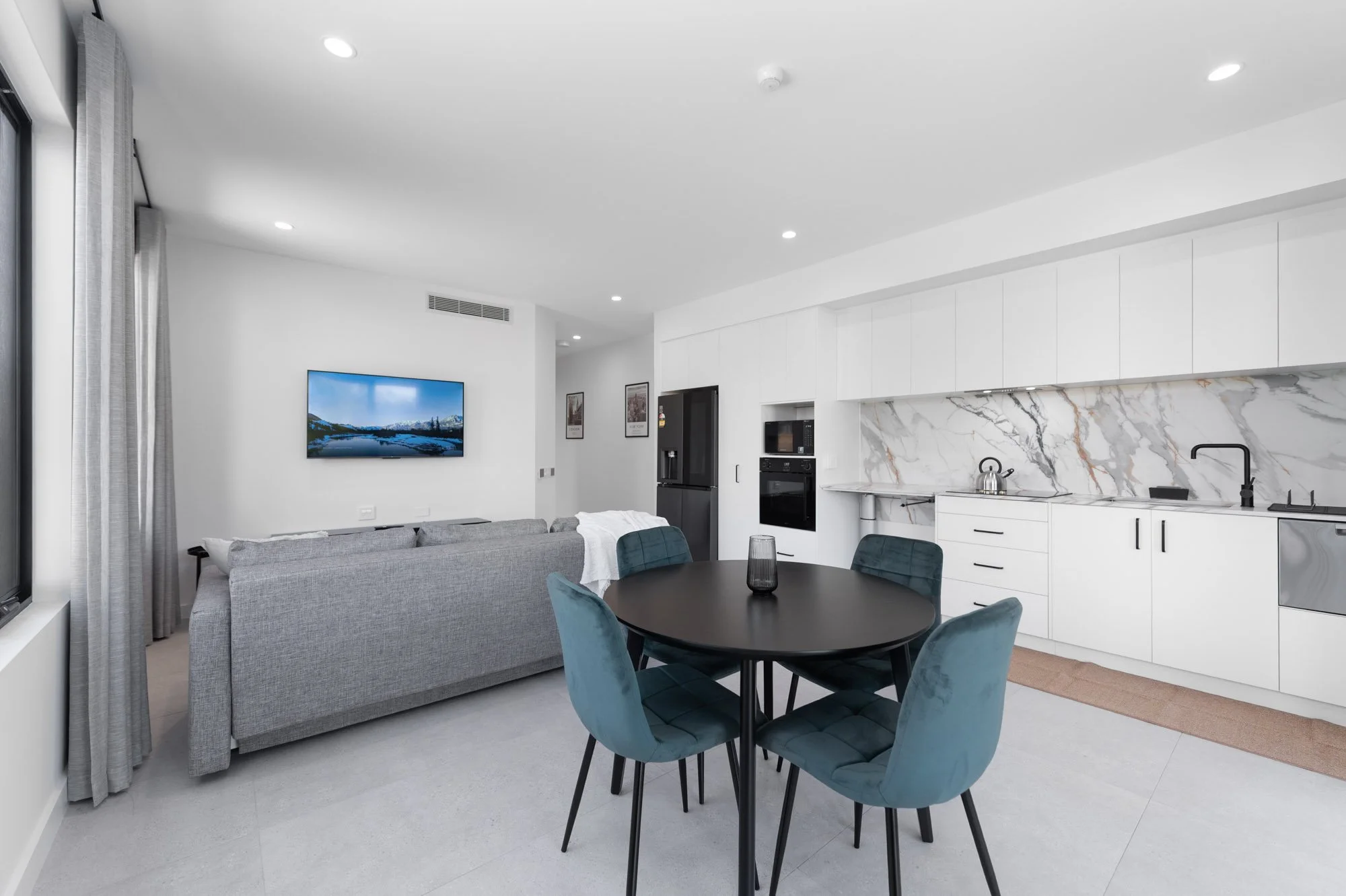 Open-concept living room and kitchen with white cabinets, gray sofa, round black dining table with four teal chairs, marble backsplash, and a wall-mounted flat-screen TV.
