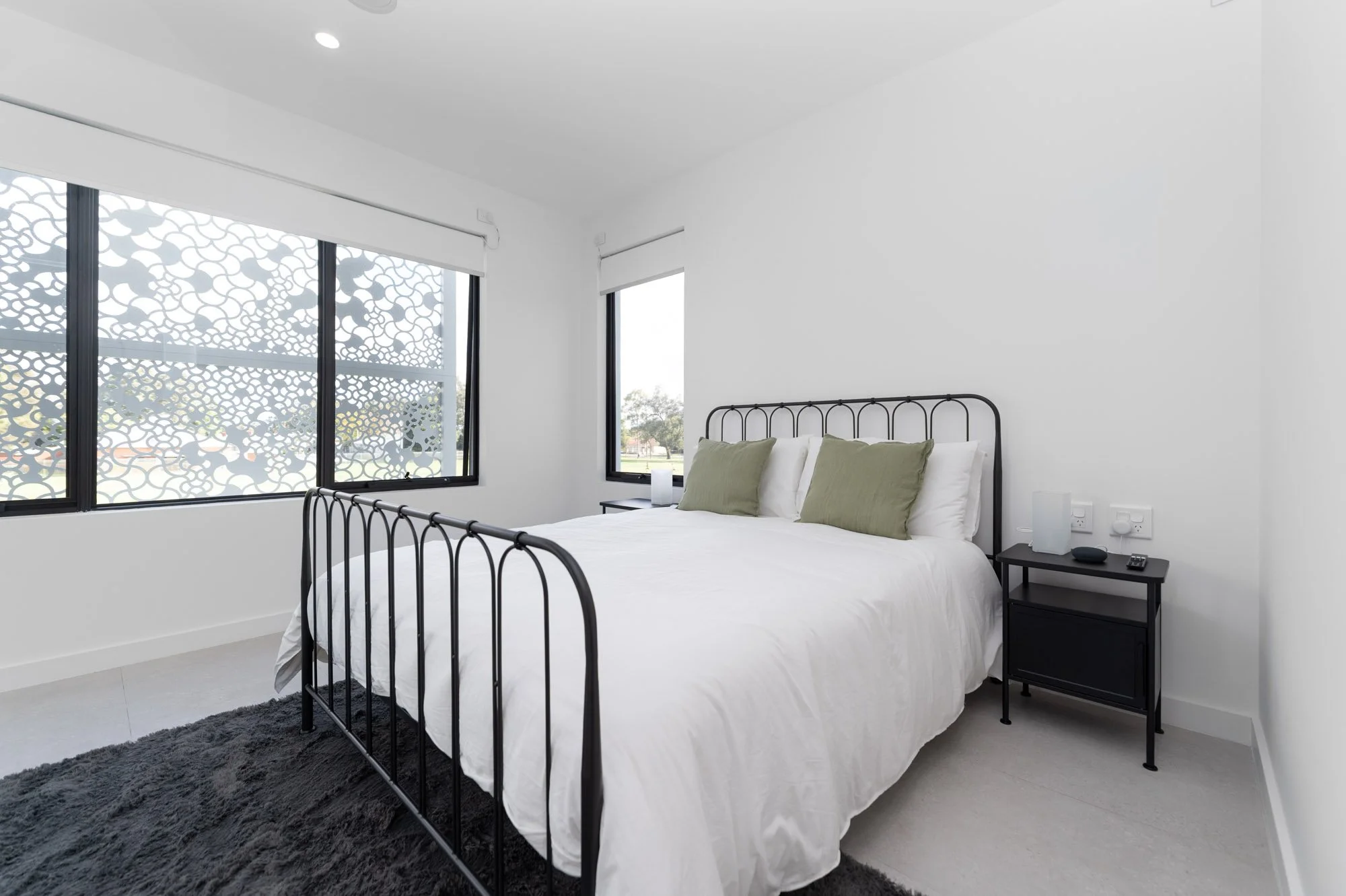 Modern bedroom with white walls, black metal bed frame, white bedding, two green pillows, black nightstand with small lamp and objects, large windows with patterned shades, and a black rug.