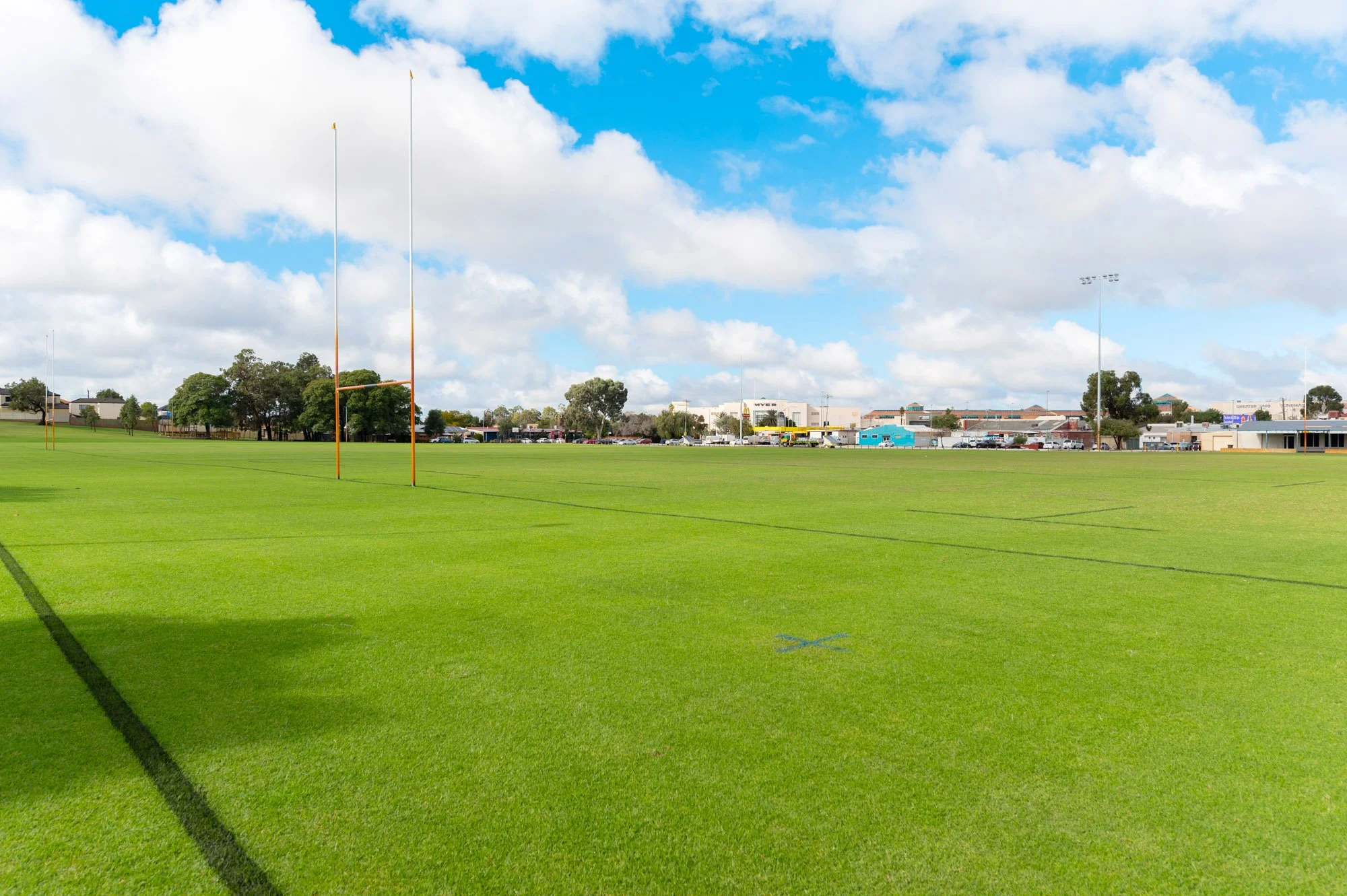 Empty green football field with goal posts under a blue sky with scattered clouds, surrounded by trees, buildings, and parking lot in the background.