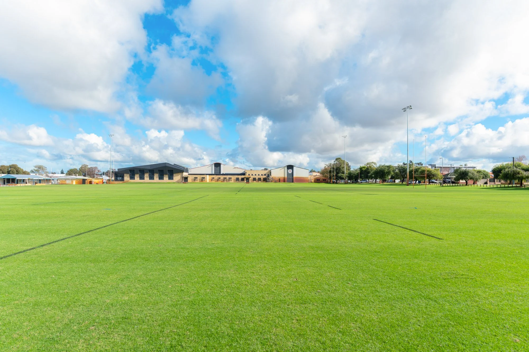 Wide grassy sports field with marked lines, school buildings in the background, along with trees and tall light poles under a partly cloudy sky.