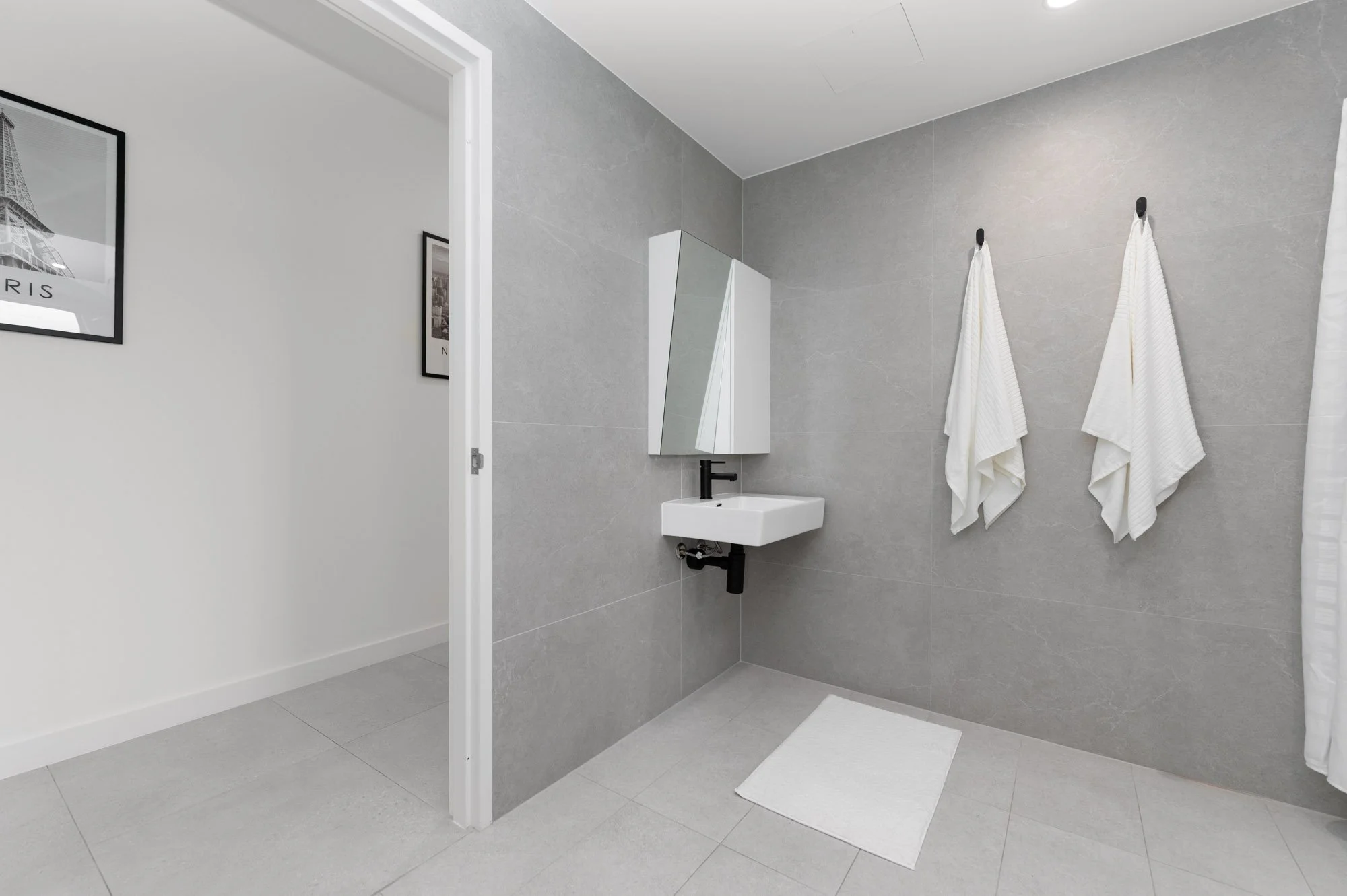 A modern bathroom with gray tiled walls and floor, a small white sink with black faucet, a mirrored cabinet, and two white towels hanging on the wall.