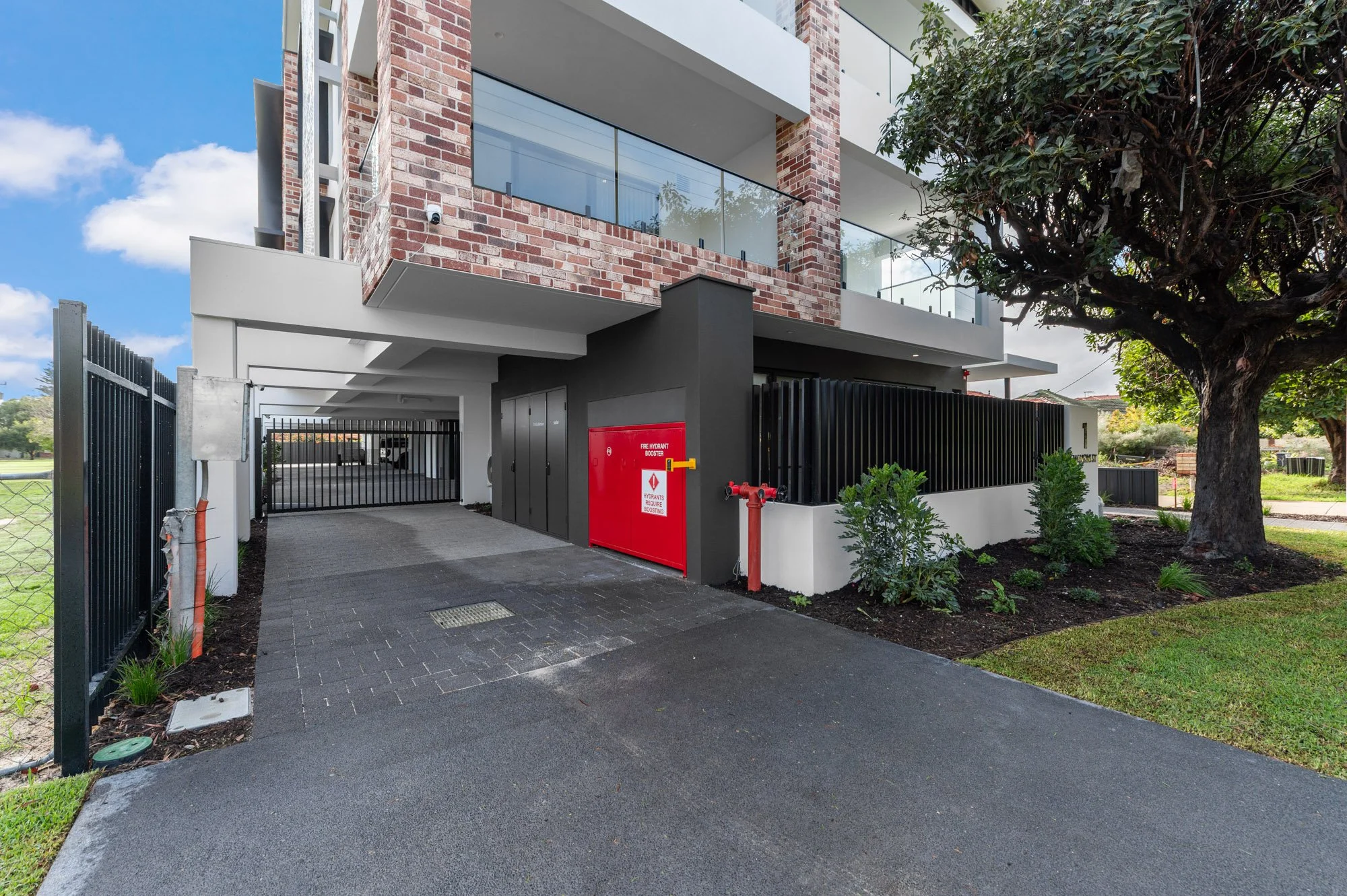 View of a modern multi-story residential building with brick and white exterior, a black gated driveway, and mature trees in a landscaped yard.