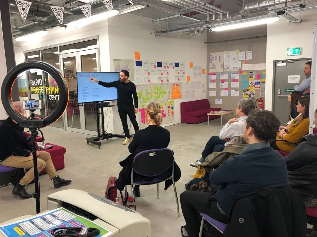 Me delivering a show and tell in the Alder Hey Innovation space. I'm pointing at a screen with an audience next to a wall of print outs and post it notes.