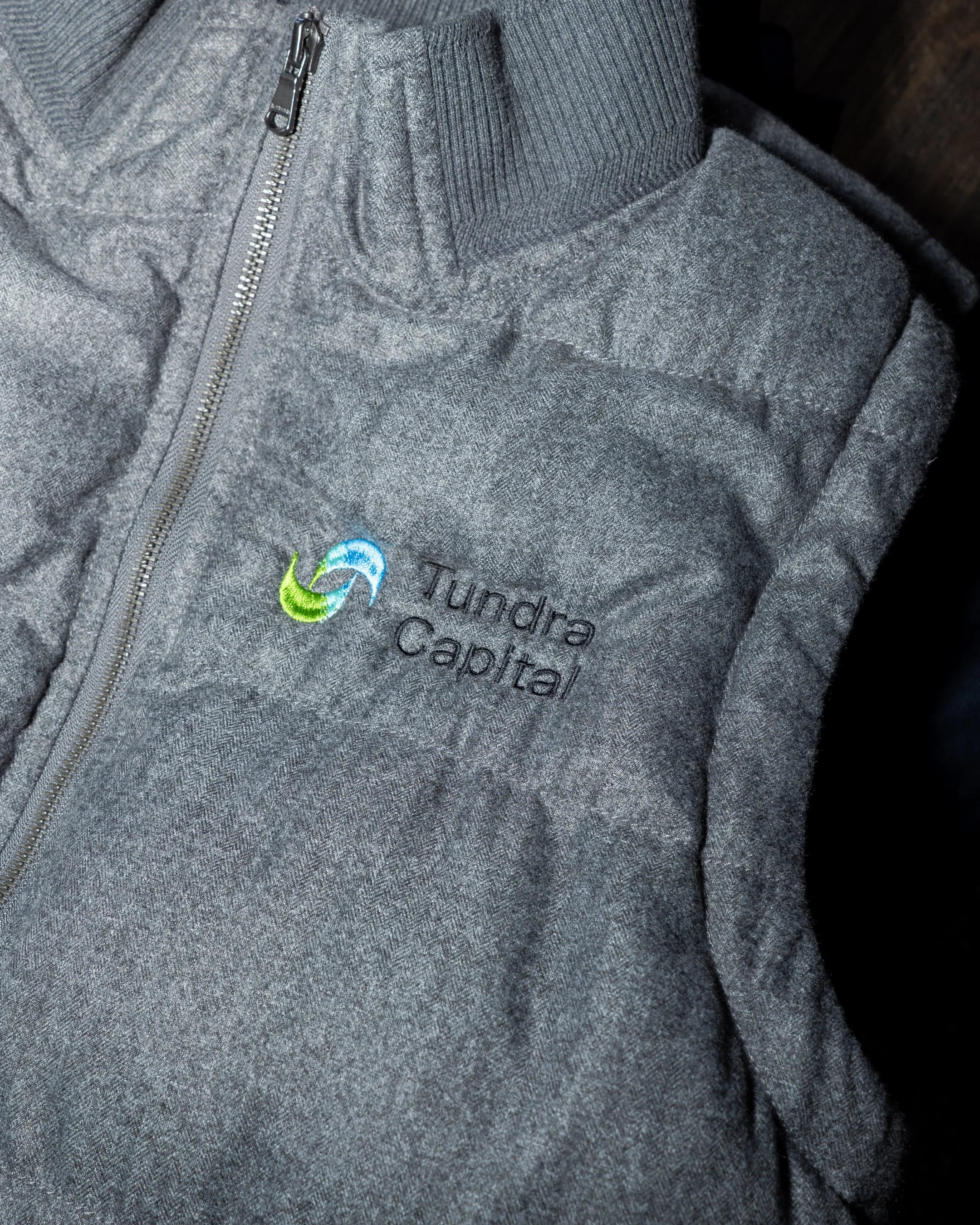 Close-up of a gray fleece jacket with a zipper, embroidered logo, and text that reads 'Tundra Capital'.