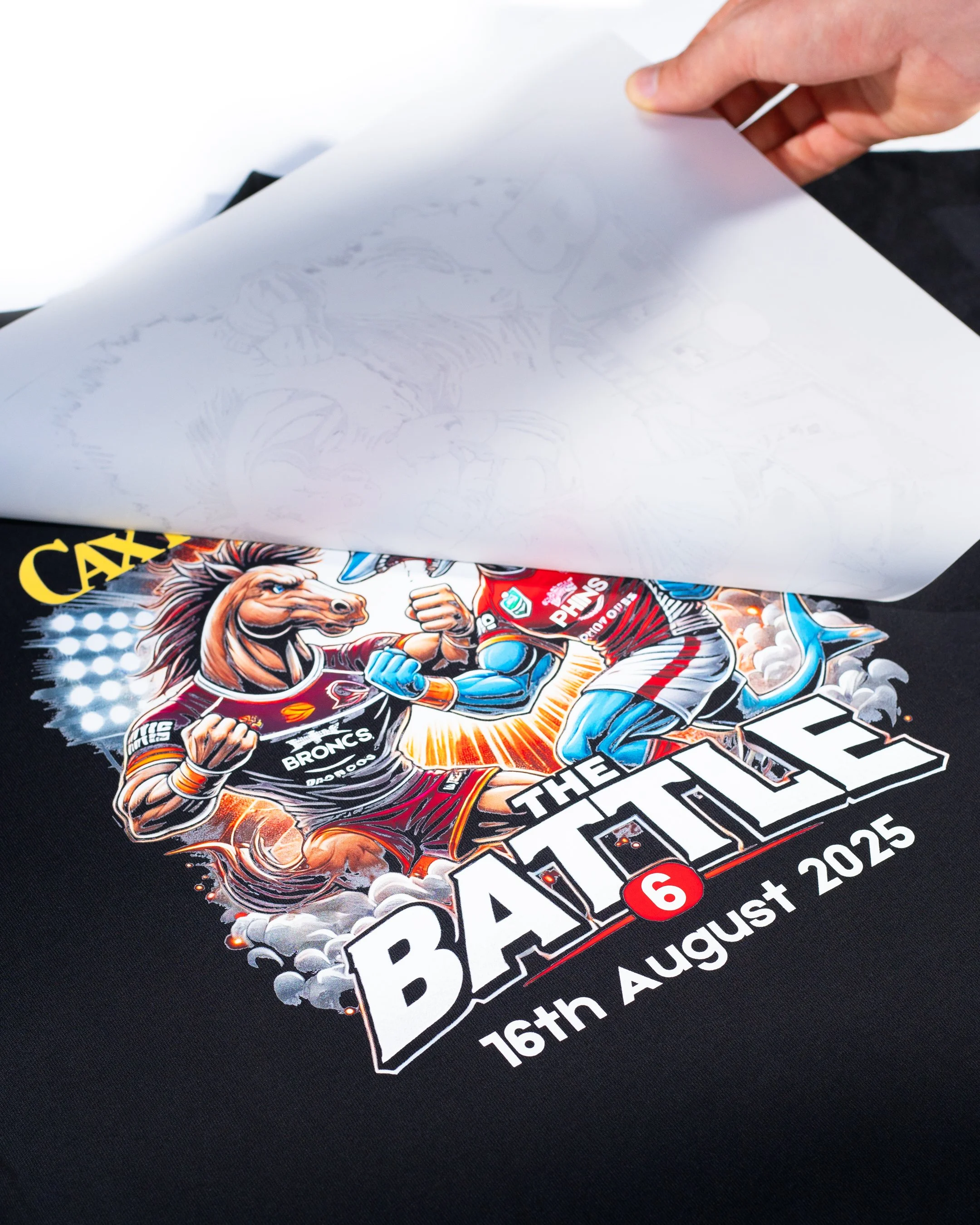 A DTF printing film being peeled on a promotional t shirt, revealing a design, featuring NRL rugby teams, the Dolphin and the Broncos, with bold text and graphics, partially visible.